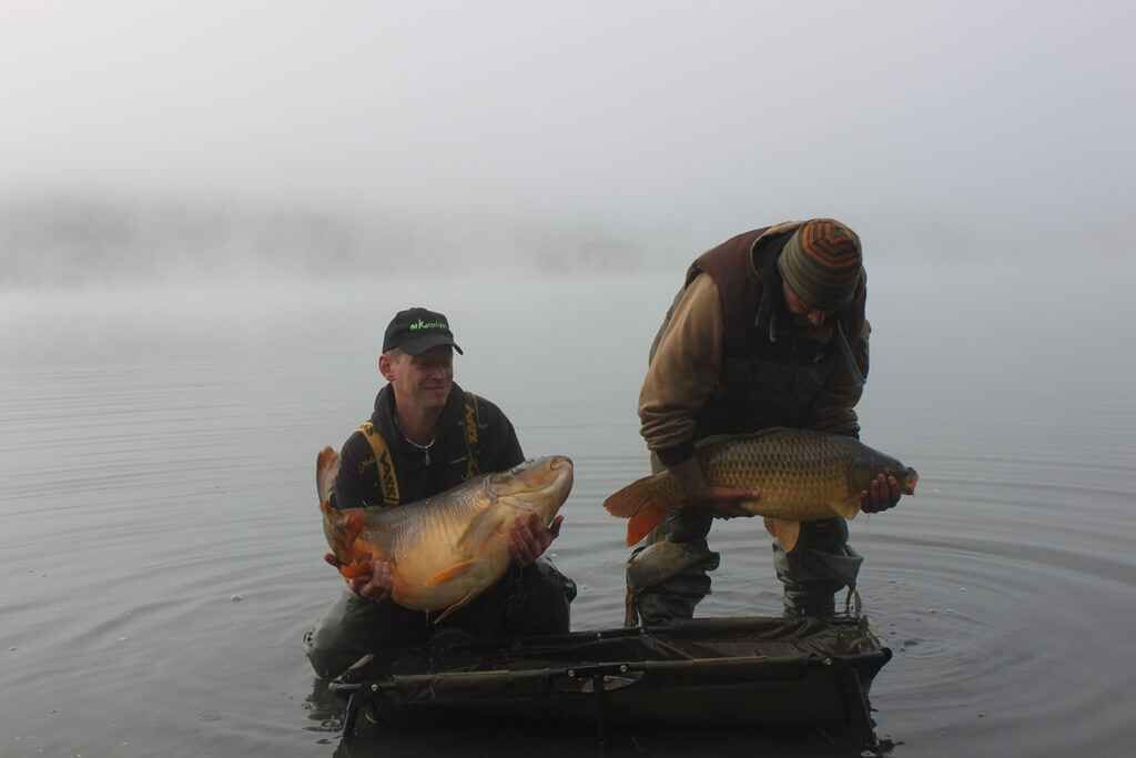 IMG_2872 Zwei Angler halten stolz große Karpfen in der nebligen Seelandschaft und präsentieren ihren Fang.