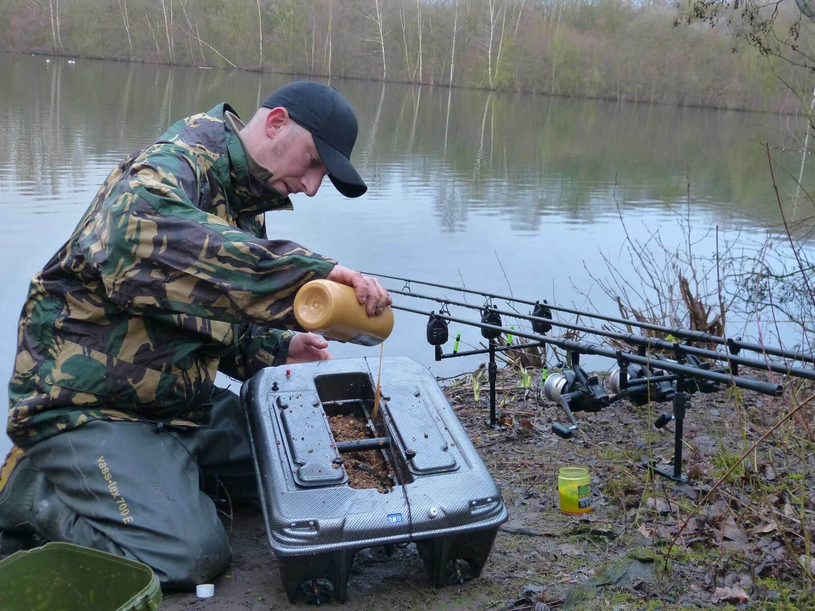 carp-madness-futterboot Angler bereitet Köder am Wasserrand vor, umgeben von Angelausrüstung und Angelruten.