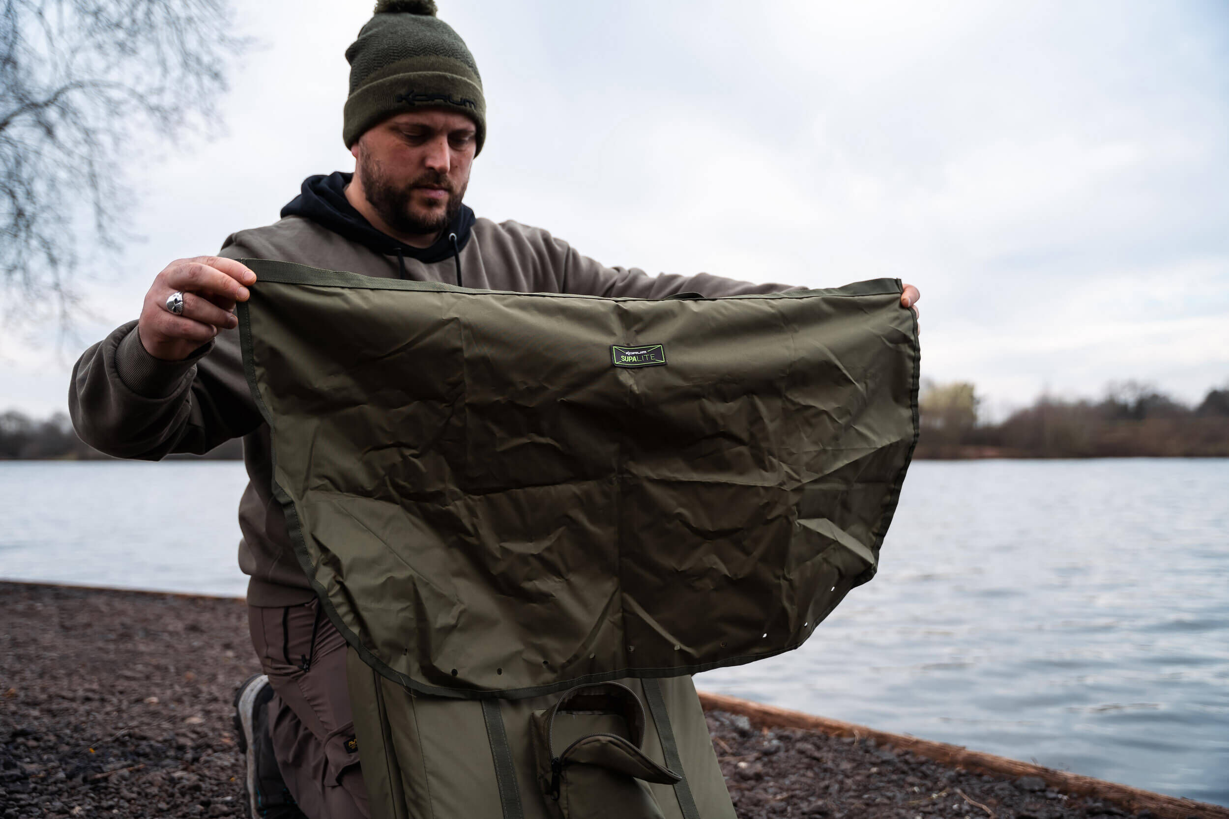 Angler inspiziert eine robuste, wasserdichte Tasche für Angelausrüstung am Seeufer.