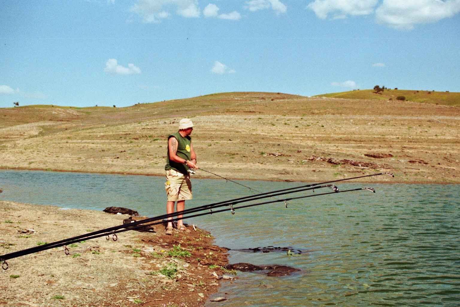 Barsche-rgern Angler angeln vom Ufer aus mit mehreren Ruten und genießen einen ruhigen Tag am Wasser.