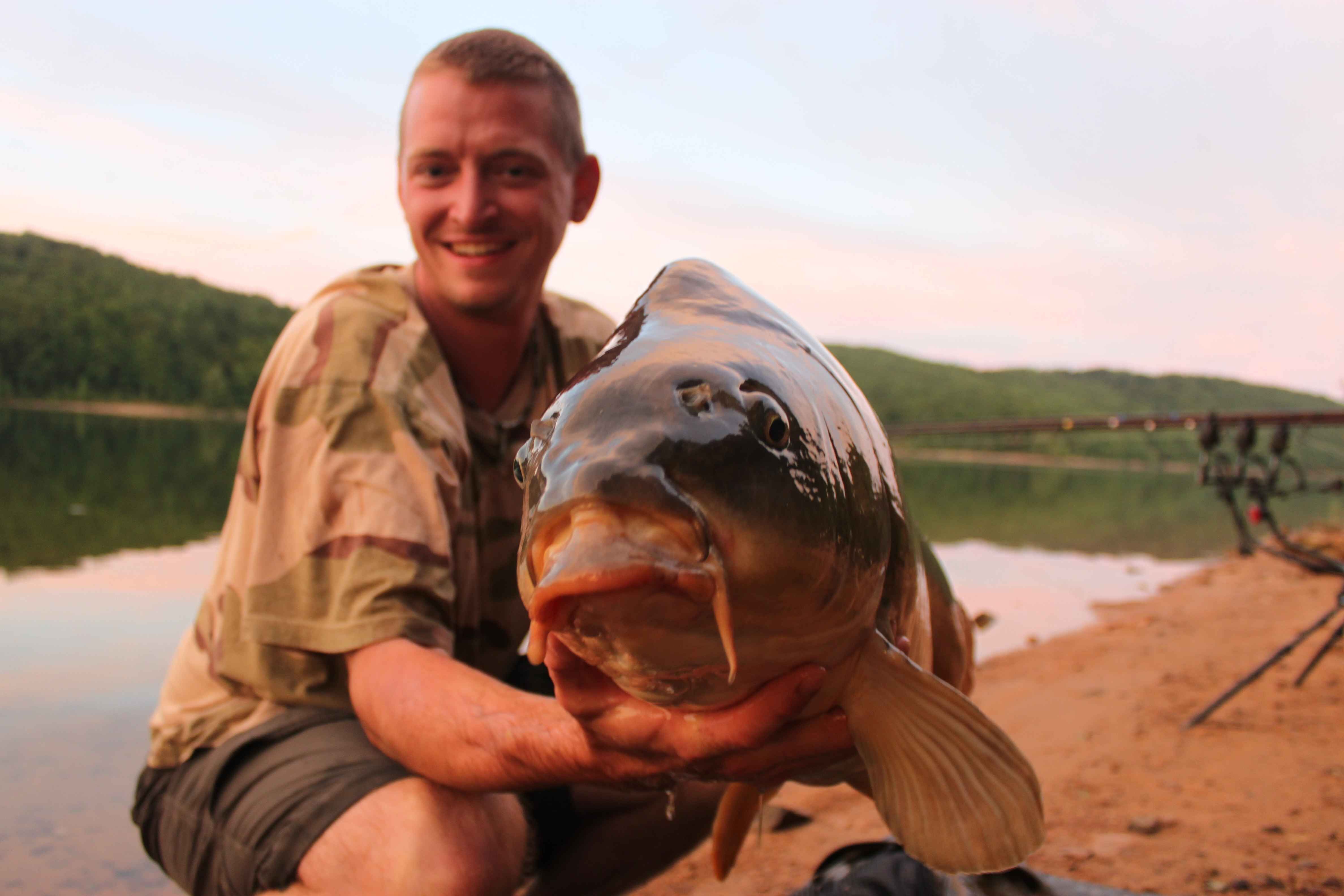 022d Ein glücklicher Angler hält stolz einen großen Karpfen in der Hand, der an einem ruhigen Seeufer gefangen wurde.