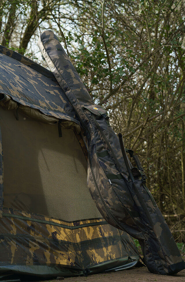 Camo hengeltas leunend tegen een bivvy voor karpervissen in een bosrijke omgeving aan het meer.