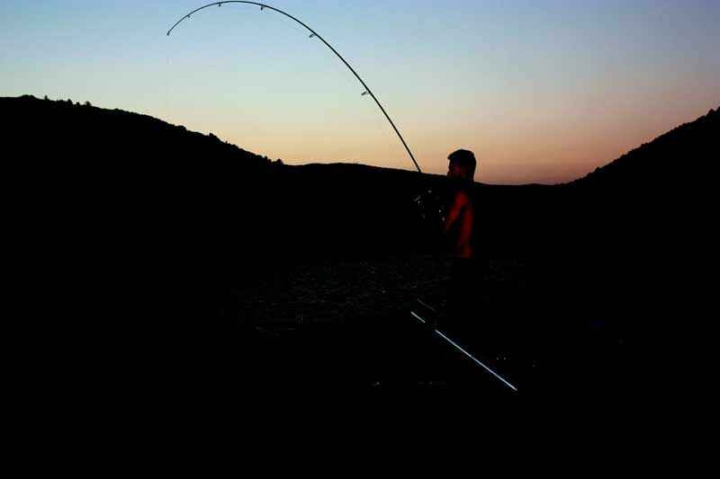 Die Geschichte der Angelrolle und Angelrute Angler angeln bei Sonnenuntergang und zeigen den Nervenkitzel des Karpfenangelns mit gebogener Rute.