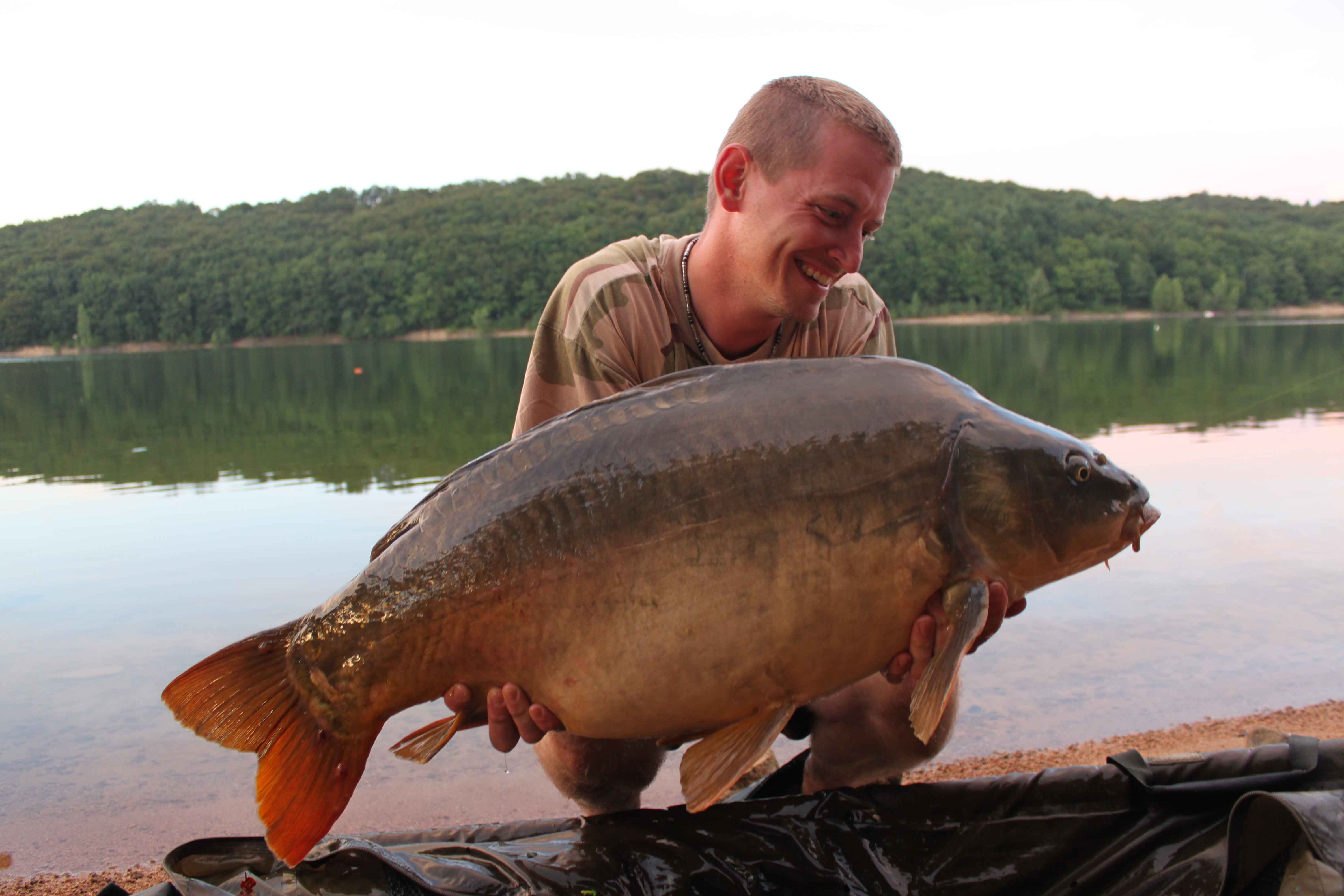 022b Ein glücklicher Angler hält stolz einen großen Karpfen in der Hand, der an einem ruhigen See gefangen wurde, und zeigt damit seinen Angelerfolg.