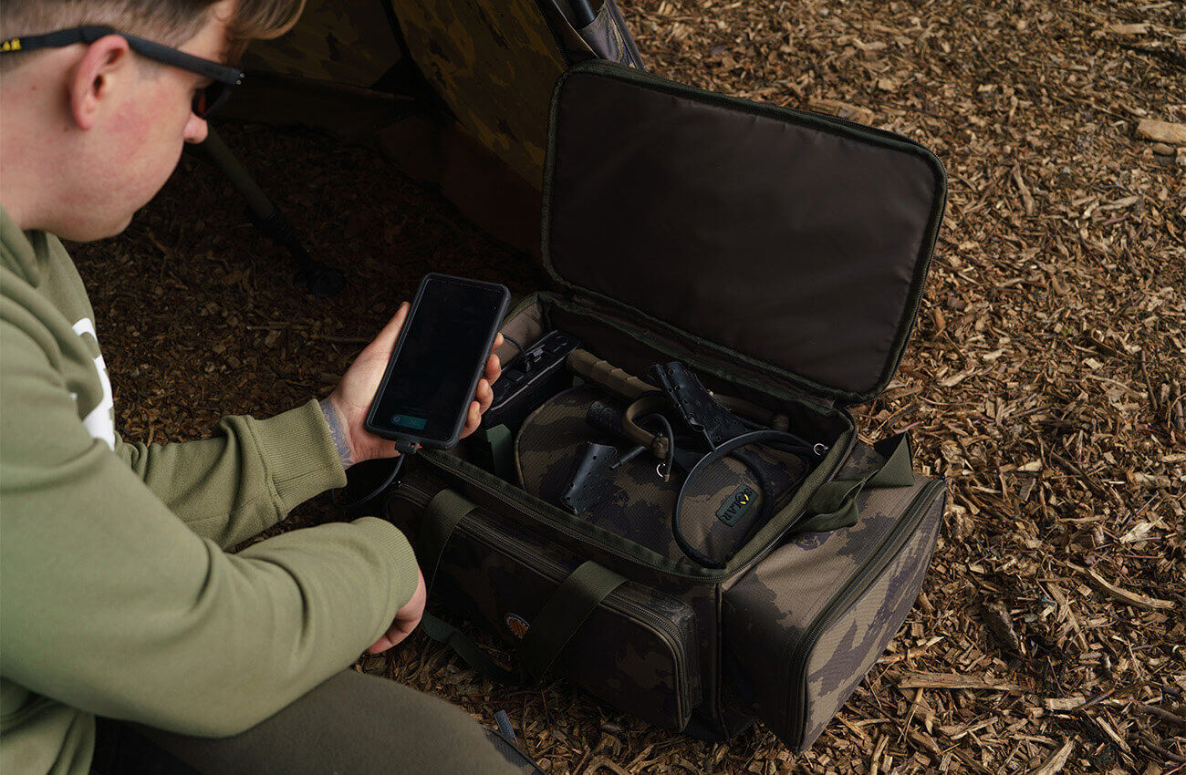 Ein Angler lädt ein Telefon neben einer Tasche mit Tarnkarpfen-Angelgerät in einem Angel-Bivvy-Zelt auf.