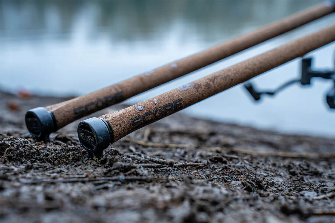 Dos cañas de pescar descansando en la orilla fangosa, listas para una aventura de pesca de carpas.