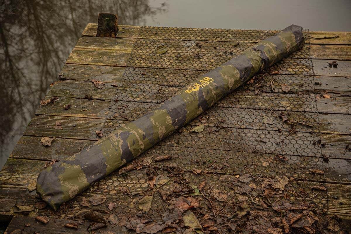 solar-CA21-brolly3 Eine Rutenhülle im Tarnmuster, die auf einem Holzsteg ruht und sich perfekt zum Schutz von Angelruten eignet.