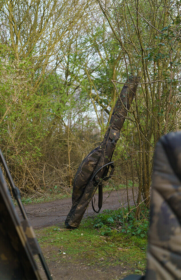 Eine Angelruten-Reisetasche im Tarnmuster lehnt an einem Baum auf einem Weg in der Nähe eines Karpfenangelplatzes.