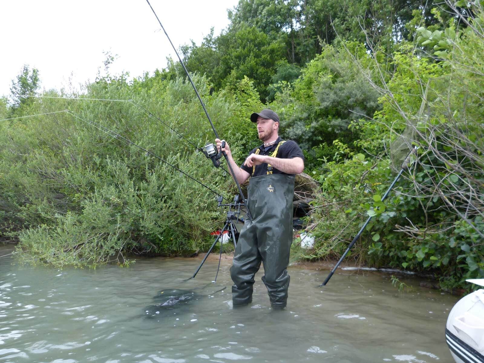 karpfenangeln-am-wasser Angler in Wathosen beim Angeln in einer üppigen, grünen Umgebung am Seeufer mit Angelausrüstung.