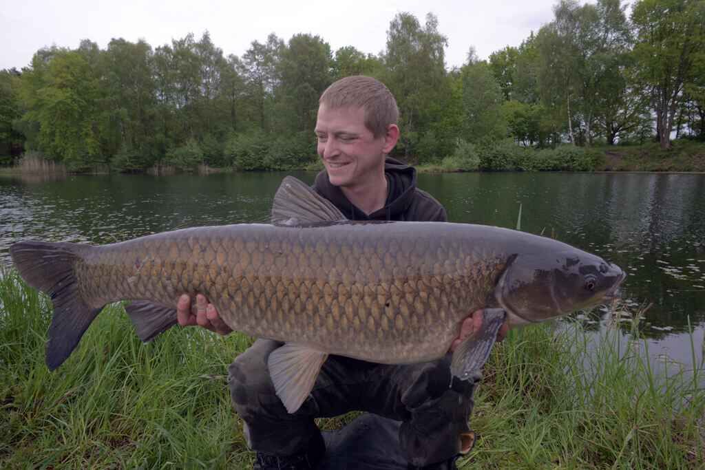 WP5_1188 Angler hält stolz einen großen Karpfen in der Hand, der an einem ruhigen See gefangen wurde, und stellt damit sein Angelkönnen unter Beweis.
