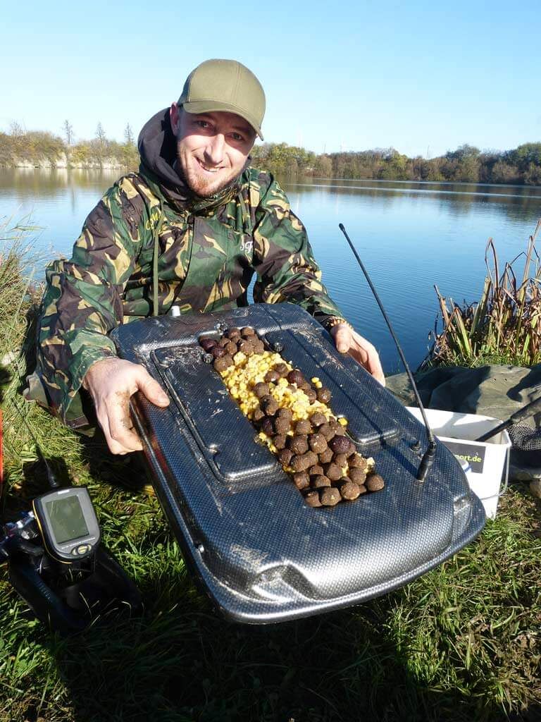 futterboot-im-einsatz Angler zeigt ein mit Karpfenködern beladenes Futterboot für erfolgreiches Angeln.