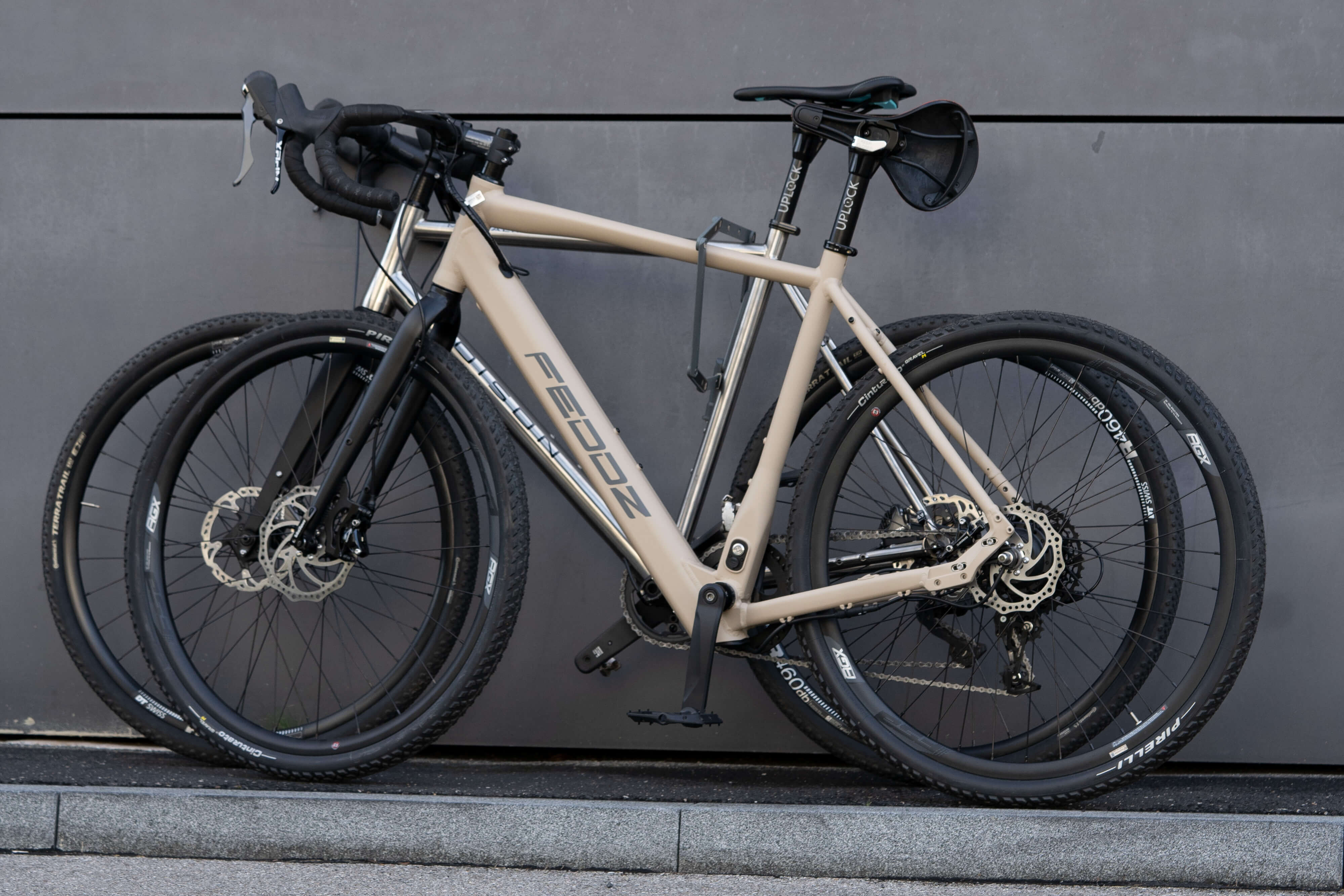Ein beiges FEDDZ-Gravelbike mit Drop-Lenker und Stollenreifen, das an einer grauen Wand lehnt.