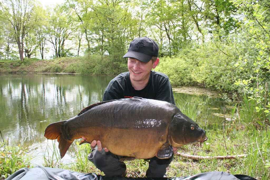 IMG_0570 Angler hält stolz einen großen Karpfen in der Hand, der an einem ruhigen Angelplatz inmitten der Natur gefangen wurde.