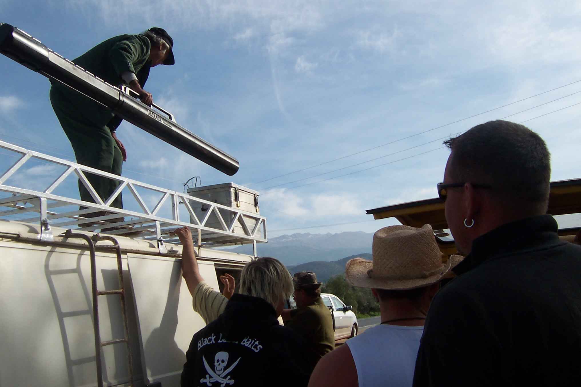 Auto-Nr2-R-ckfahrt-BeneMilla-Casablanca-3 Angler versammeln sich, während ein Mann bei klarem Himmel auf dem Dach eines Fahrzeugs Angelausrüstung vorbereitet.